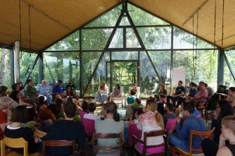 People sitting in concentric circles in a large tent with glass sides, sunlight coming in and trees out the window